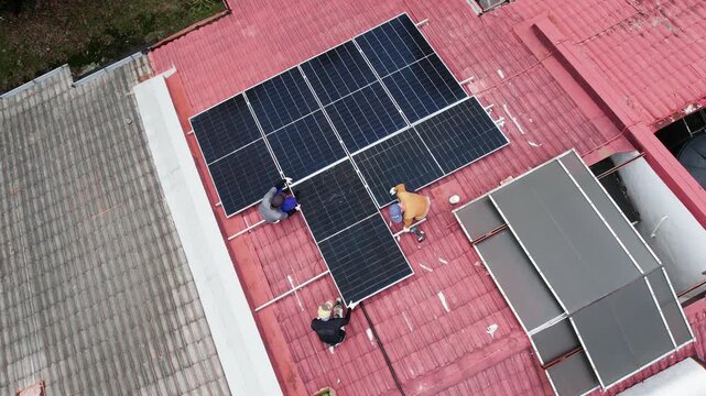 Aerial view of unrecognizable workers installing solar photovoltaic or PV panels onto red rooftop of residential house in Malaysia