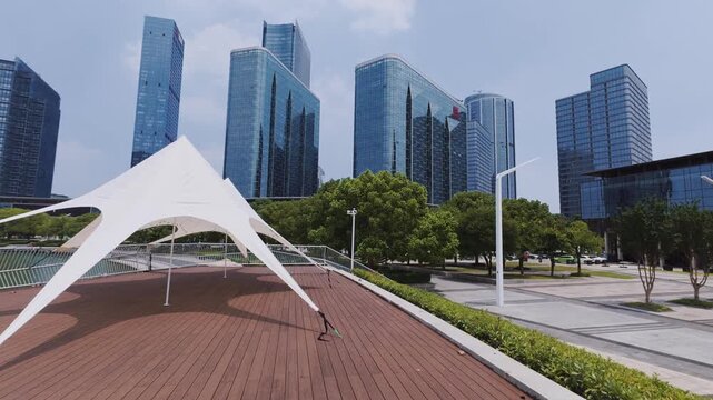 A modern waterfront promenade with a tensile shade canopy, set against a backdrop of tall glass skyscrapers in an urban cityscape.