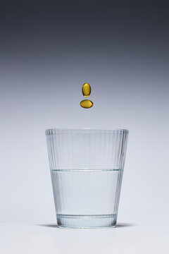 Falling yellow vitamin capsules above a glass of water on grey background