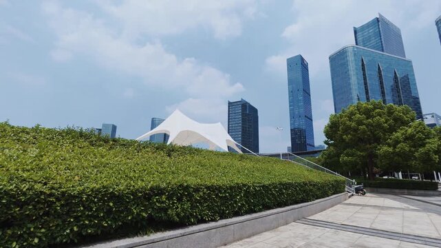 A modern urban plaza with green landscaped slopes, surrounded by glass skyscrapers under a clear blue sky.
