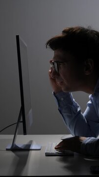 Vertical video young man wearing glasses experiencing fatigue and eye strain from prolonged screen time in a dark office, highlighting concepts of overwork, stress, and technology addiction