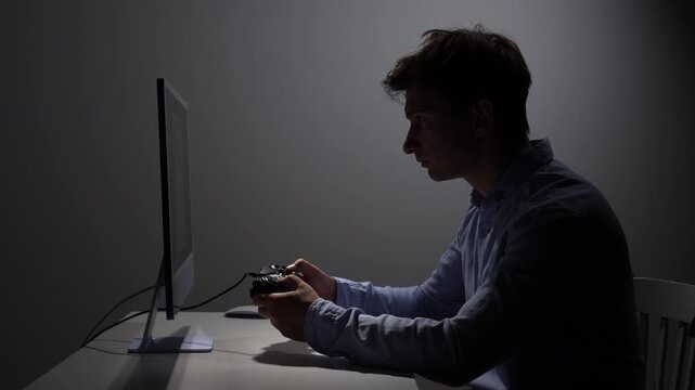 Young man concentrating on a computer screen, holding a game controller while gaming in a dimly lit room, depicting a modern leisure activity and digital entertainment experience