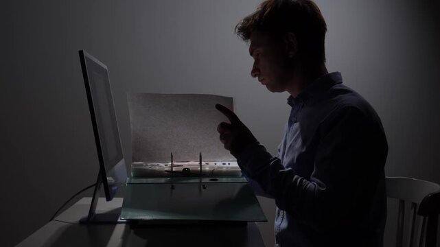 Man in a dimly lit office diligently working through documents in a binder next to a computer, focusing on his tasks during extended hours