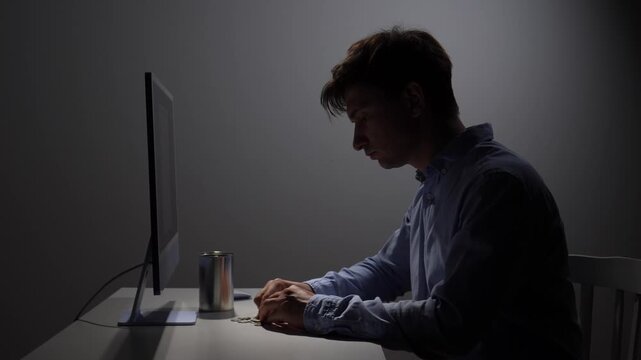 Man in a dark room intensely counting coins from a container, sitting at a desk with a computer monitor, symbolizing financial stress, debt, and economic hardship during a difficult period