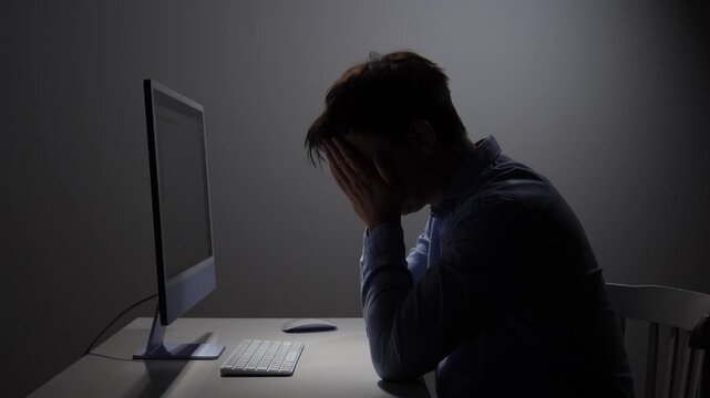 Man covering his face with hands, showing extreme fatigue and frustration while working late at night in a dim office setting, dealing with deadlines and overwork
