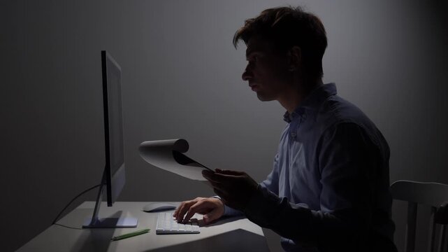 Overworked man focusing intently on a document and typing on a computer keyboard, working late at night in a dimly lit room, symbolizing stress, deadlines, and remote work