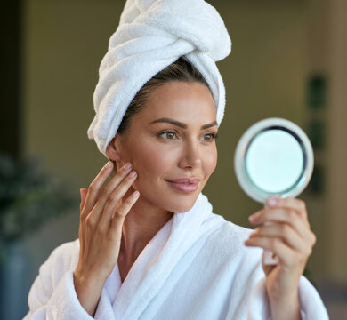 Woman in bathrobe and towel turban looking into round mirror while touching face.