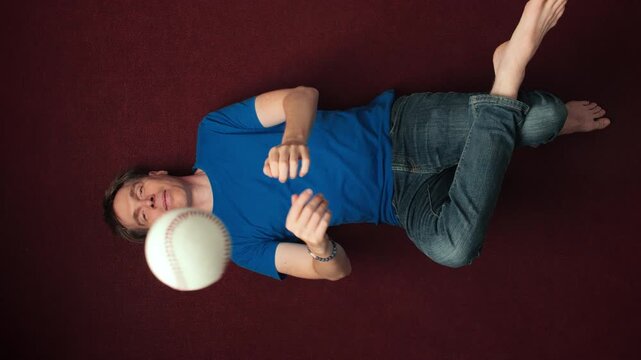 Young man lying on the floor and throwing and catching a white ball in boredom. Overhead shot with rotation.
