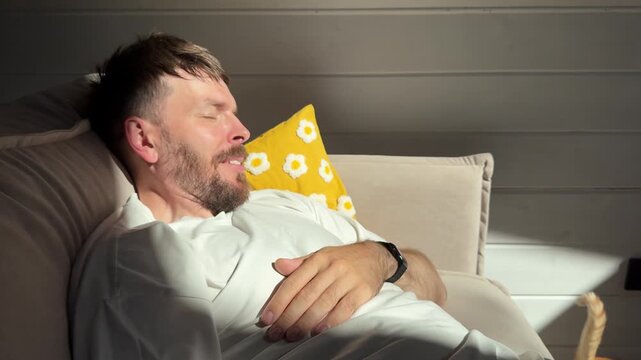 A young man with a beard, who looks like a Scandinavian, is playing with a little red-haired kitten on the couch; and the kitten jumps down onto the floor