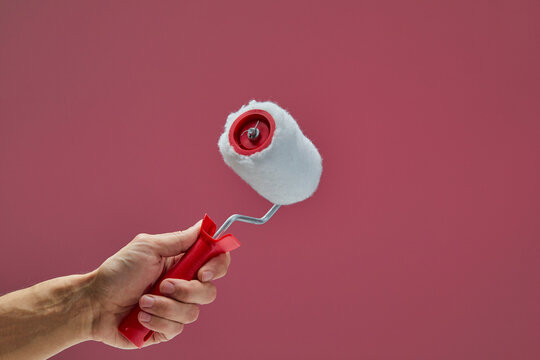 Red paint roller with white fluffy cover, gripping hand preparing painting surface against bright pink background