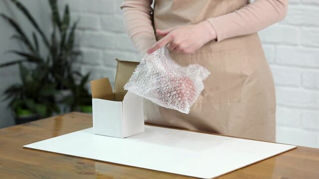 Woman packing fragile ceramic teapot in bubble wrap for safe cardboard box shipping