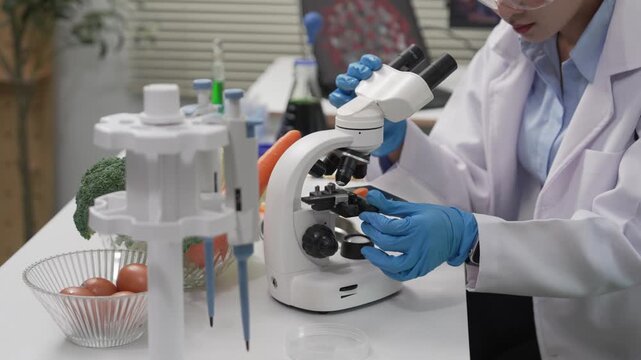 An Asian female food scientist wearing safety glasses and gloves holds fresh broccoli and carrots, inspecting their quality for pesticide residues or chemical contaminants in a food research laborator