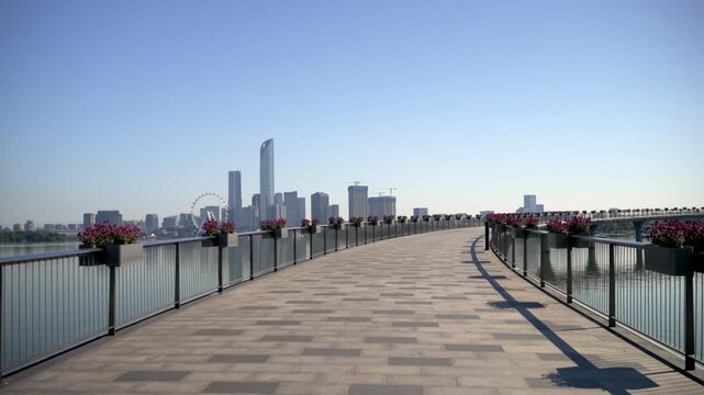 A curved waterfront promenade with flower planters overlooks a modern city skyline under a clear blue sky.