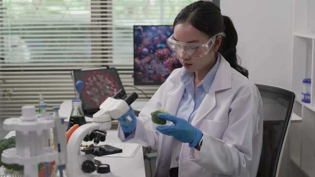 An Asian female food scientist wearing safety glasses and gloves holds fresh broccoli and carrots, inspecting their quality for pesticide residues or chemical contaminants in a food research laborator