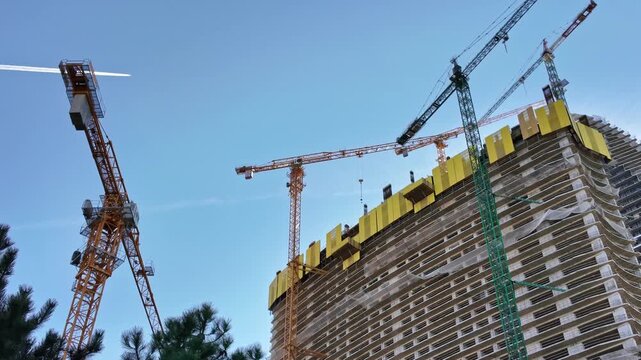 Multiple tower cranes operate above a rising high rise building construction site against clear blue sky.