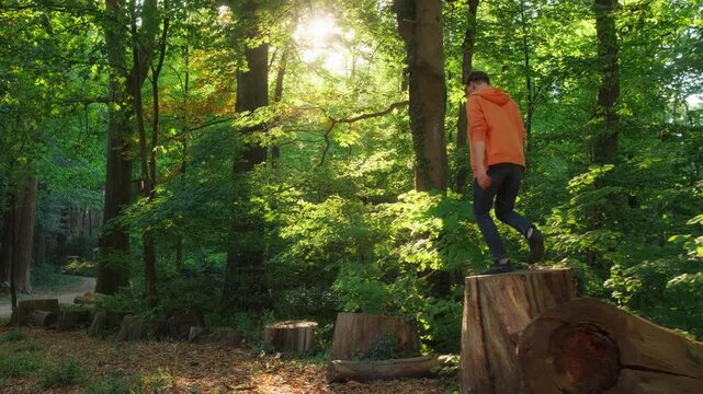 Young man playfully jumping on logs in a sunny lush green forest, with the beautiful sun in the background