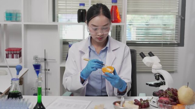 Asian female food scientist wearing lab coat, safety glasses, and gloves examining a fruit sample under a microscope and recording data on a clipboard in a modern microbiology food safety laboratory.