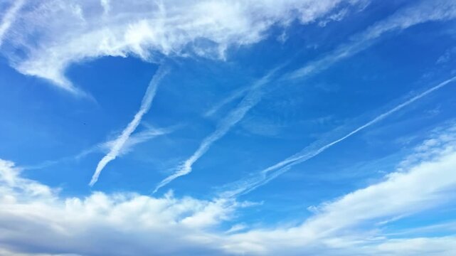 White airplane contrails crisscross a bright blue sky with soft clouds drifting below in daylight.