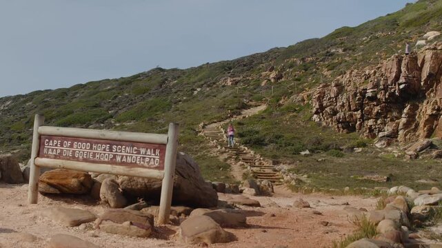Visitor walks uphill beside Cape of Good Hope Scenic Walk sign, following rocky trail through low coastal vegetation and cliffs. Travel footage conveys outdoor exploration and scenic hiking.