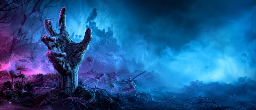 A zombie hand reaching out of the grave on a spooky Halloween night; tombstone, thick fog, and blue background (banner format)