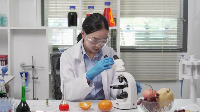 Asian female food scientist wearing lab coat, safety glasses, and gloves examining a fruit sample under a microscope and recording data on a clipboard in a modern microbiology food safety laboratory.