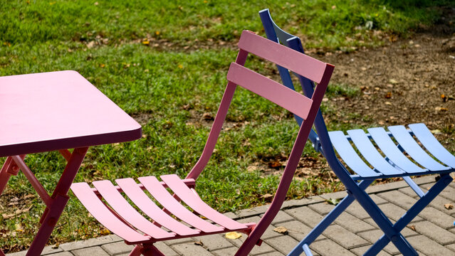 Colorful garden furniture inviting outdoor relaxation and leisure. Featuring a pink table and chair alongside a blue chair on a paved patio with green grass in the background