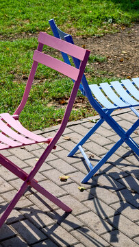 Two colorful folding metal chairs. One pink and one blue. Standing on a paved surface next to green grass. Casting long shadows under sunlight