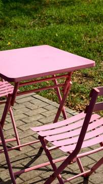 Pink metal outdoor bistro set with a square folding table and two matching chairs. Placed on a paved patio beside a green lawn