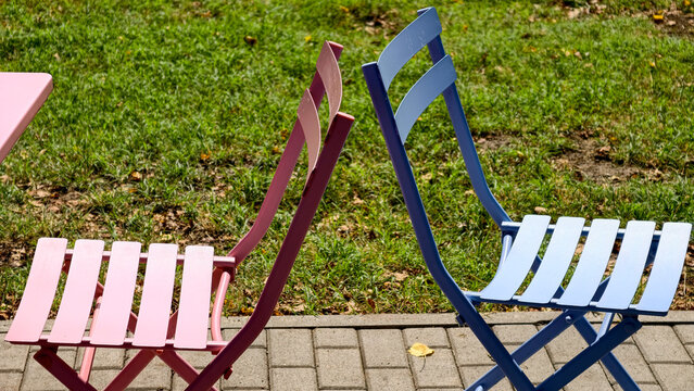 Pink and blue metal folding chairs standing back to back on a paved area beside green grass, symbolizing separation, disagreement, or contrasting viewpoints