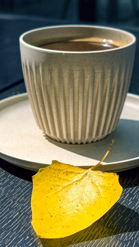 Warm coffee in a textured mug sits beside a bright yellow fallen leaf on an outdoor cafe table, embodying the peaceful and refreshing start to an autumn day