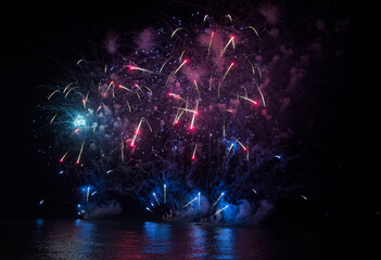 Fototapeta premium Vibrant fireworks burst in the night sky, illuminating a grand building and its colorful reflections on the calm water below.