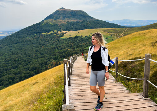 blonde woman hiking on wooden walkway in green forested hills with rope railings, serene mountain landscape overhead, peaceful nature adventure travel scene, The Puy de Dome in Auvergne