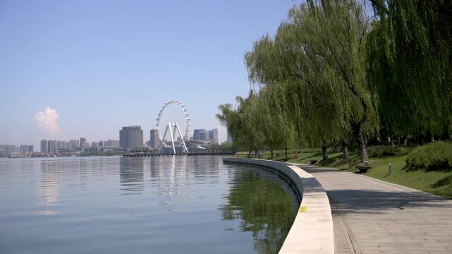 A calm waterfront walkway with willow trees, distant urban buildings, and a ferris wheel under a clear sky.