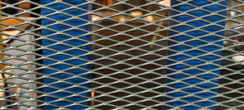 Steel mesh grid with diamond-shaped pattern in front of blue corrugated surfaces and machinery elements. Industrial close-up emphasizing reflective metal texture and mechanical background.