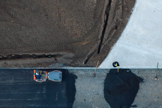 Belgrade, Serbia - 14 November 2025: Aerial view of workers laying new asphalt during road construction, with a steamroller compacting the surface and a worker using a shovel near a pile of gravel.
