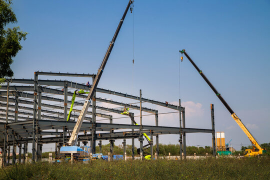 Large Steel-Frame Warehouse Structure Under Construction with Crane and Worker,Construction Workers on Cherry Picker Positioning High-Beams for New Facility