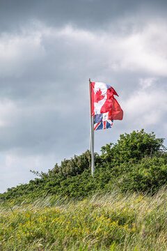 National flags of Canada and the United Kingdom flying together on a flagpole on a green hill. Allied memorial site at Juno Beach in Normandy under a dramatic cloudy sky.