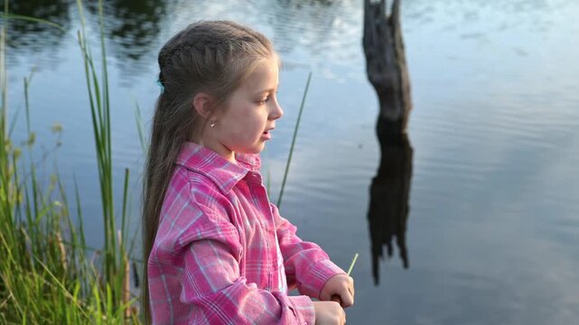 Young girl plays with cattail beside calm lake, holding stem and brown seed head near her face. Child explores nature through outdoor play, summer recreation, curiosity, and lakeside background.
