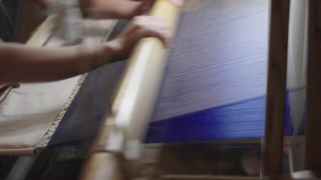 A person's hand operates a wooden traditional loom to weave fabric, demonstrating manual textile craft work.