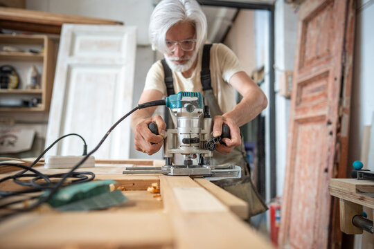 Professional carpenter working on restoration of antique wooden door in restoration workshop, craftsman using electric plunge router machine, processing door surface at workbench with instrument.