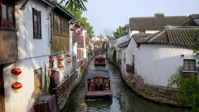 A narrow canal in a traditional water town, with tour boats navigating between white-walled old buildings adorned with red lanterns.