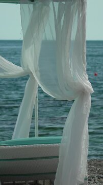 Ocean breeze moves translucent fabric canopy over a comfortable wooden sunbed near turquoise waves. Stationary eye-level tripod shot.