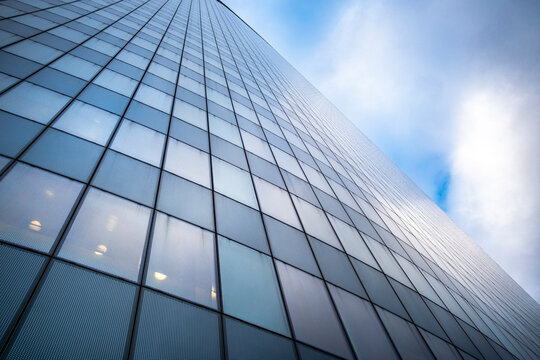 A modern building rises in upward view as glass facade and windows catch blue sky and clouds, giving the architecture a clean reflective presence