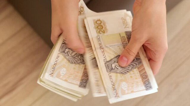 Hands counting a large stack of Polish zloty banknotes on a wooden floor from a top-down perspective, illustrating personal finance, savings and cash management. High quality 4k footage