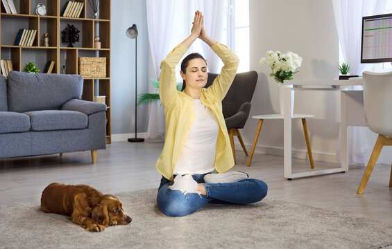 Woman and dog practice meditation at home. In a calm living room, she sits with hands raised in yoga prayer while a relaxed cocker spaniel rests nearby. Mindfulness, calm, and pet bonding.