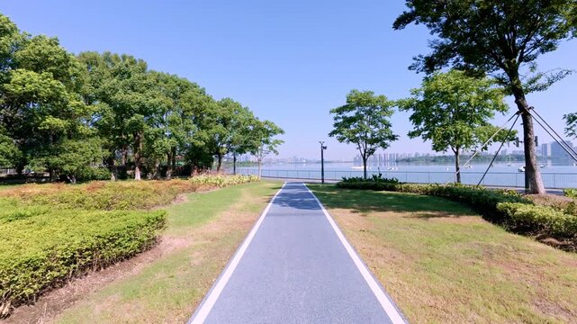 A paved lakeside path extends toward calm water, flanked by green trees and lawns under a clear sunny sky, forming a tranquil outdoor scenic space.