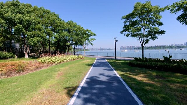 A clear sunny day at a lakeside park, with a paved path flanked by grass and trees leading to calm water.