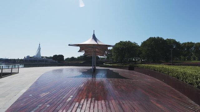 A sunlit outdoor waterfront plaza features a central canopy, wooden deck, and landscaped greenery under a clear sky.