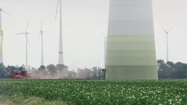Wind turbines rise above flowering crop field as agricultural machinery works through dusty farmland. Rural scene combines renewable energy, farming, harvest activity, and sustainable land use