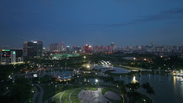 Aerial night view of Hoa Binh Peace Park in Hanoi, featuring a tranquil lake, illuminated pathways, landscaped greenery, and the surrounding city skyline.